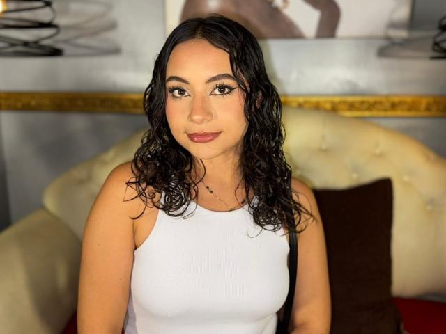 BiancaB a les cheveux noirs et bouclés, elle porte un débardeur blanc qui révèle ses petites seins, elle a les yeux marron et un léger sourire, et elle est assise sur un canapé crème..