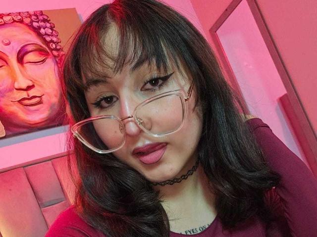 MaddieLesYoux with glasses, dark wavy hair, and red lipstick, tongue slightly out, wearing a black choker, in a pink-lit room, Buddha painting in background.