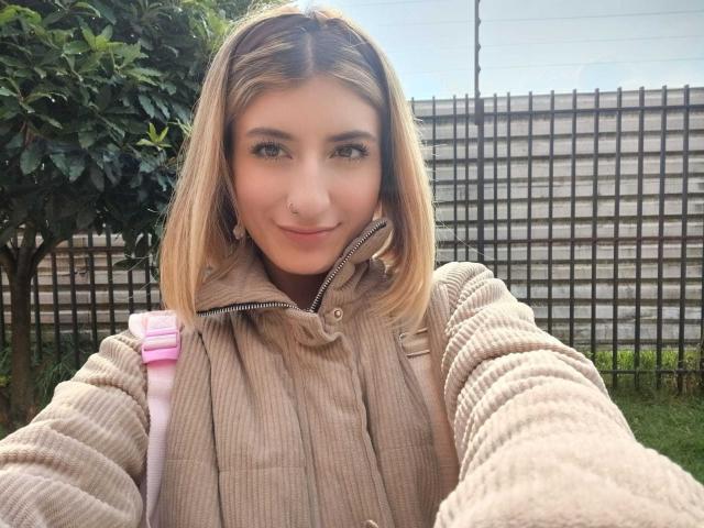 Kaiix has blonde hair, green eyes, and a cute nose ring. She's wearing a beige ribbed jacket with a pink strap. Her small tits are slightly visible.