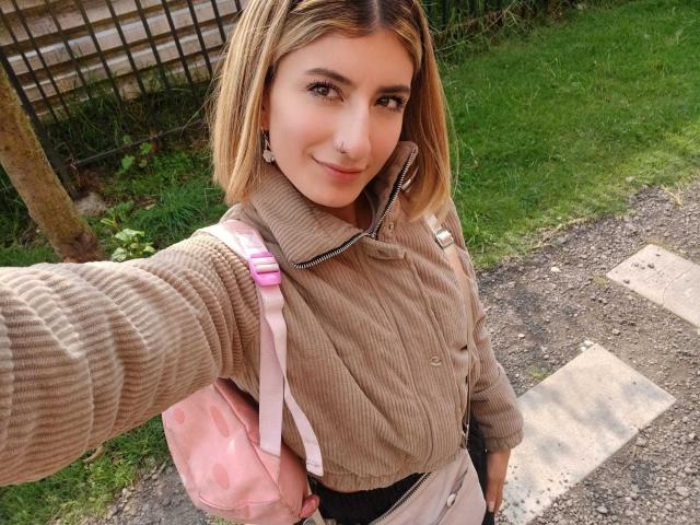 Kaiix with blonde bob, smiling, wearing a beige ribbed shirt, pink polka-dot backpack, black pants, and small earrings, taking a selfie outside. Her tits are not visible.
