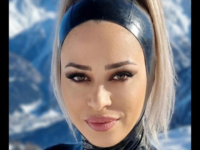 Close-up of GloriaMelek with light skin and blonde hair in a black latex headband. She has thick, arched eyebrows, long, dark eyelashes, and pink lips. Her eyes are slightly downward, giving a subtle, confident expression. The background is blurred, featuring blue and white snowy textures. Her makeup is precise, emphasizing her sharp features. The latex headband contrasts with her smooth skin and blonde hair, adding a sleek, futuristic element.
