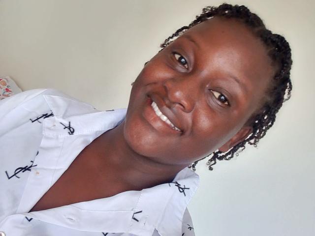 PinkishPussy has dark brown skin, small white teeth, and short braided hair. She's wearing a white shirt with black patterns, smiling at the camera.