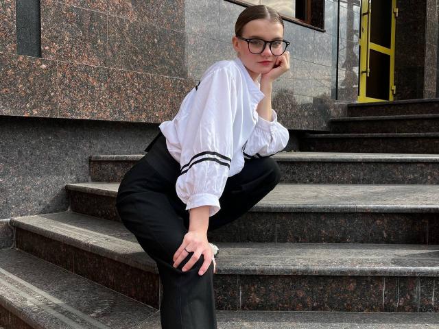 YaekoBordges, with glasses, white blouse, black pants, and tied-up brown hair, sits on marble steps, resting chin on hand, looking down, with one knee raised.