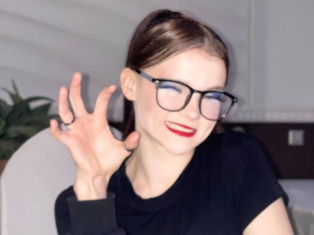 YaekoBordges met een bril, rode lippenstift en een zwart shirt, die haar rechterhand omhoog steekt, lachend met gesloten ogen, bruin haar in een paardenstaart..