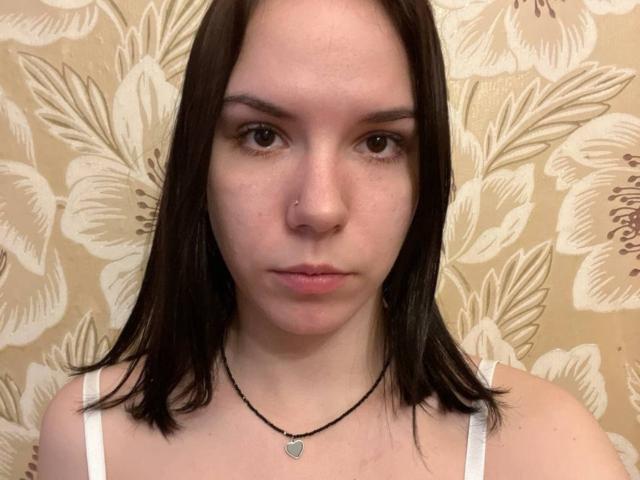 MagdalenaMurchie with straight dark brown hair, white tank top, black necklace with heart pendant, nose ring, and serious expression, showing her face and upper chest.