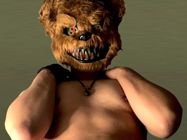 AdoniBaskerville with a bear mask has a muscular, shirtless upper body. The mask has brown, fuzzy texture, exaggerated teeth with red and green eyes. AdoniBaskerville's hands are raised, gripping the mask's edges. A black necklace with a small pendant hangs around their neck. The mask's eyes are intense and the teeth are sharp. The lighting is harsh, emphasizing the mask's details and AdoniBaskerville's toned chest. The image is digital with a slightly grainy texture.