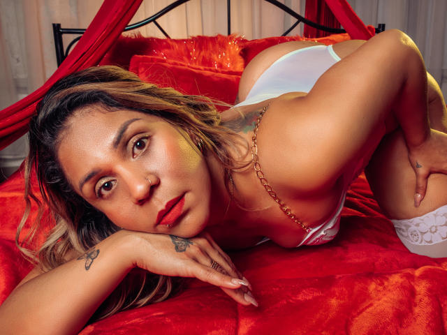 ManyHawkey è sdraiata su un letto rosso, indossa delle mutandine bianche e una collana, è a quattro zampe, ha un tatuaggio sulla mano e un piercing al naso, e guarda verso la telecamera..