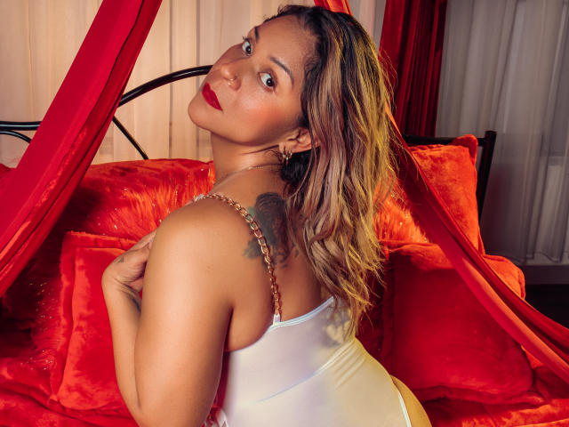ManyHawkey with red lipstick, wavy blonde hair, white tank top, revealing her back tattoo, sitting on red velvet pillows, looking over her shoulder, showing her ass.