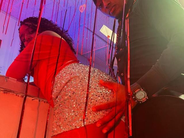LilithAndAsmodeo in a red top and sparkly ass, bent over, being grabbed by LilithAndAsmodeo's hand, wearing a watch, in a red-lit, hanging-bead-filled scene.