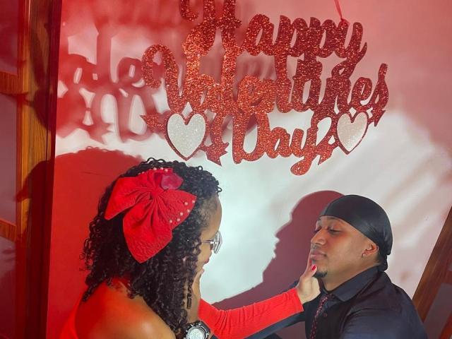 LilithAndAsmodeo with curly black hair and red bow, being gently touched by LilithAndAsmodeo in black cap and shirt, standing in red light, looking at each other.