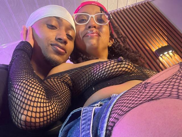 LilithAndAsmodeo, black fishnet top, small breasts, wearing denim shorts, pink hat, glasses, and white headband, sitting close to LilithAndAsmodeo with a beard, white headband, and relaxed expression.