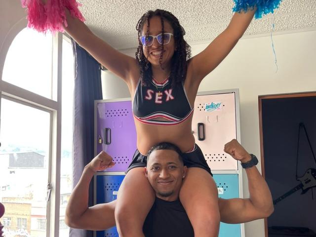 LilithAndAsmodeo, a black woman with curly hair and glasses, is sitting on a black man's shoulders, wearing a "SEX" crop top and shorts, with pink and blue pom-poms.