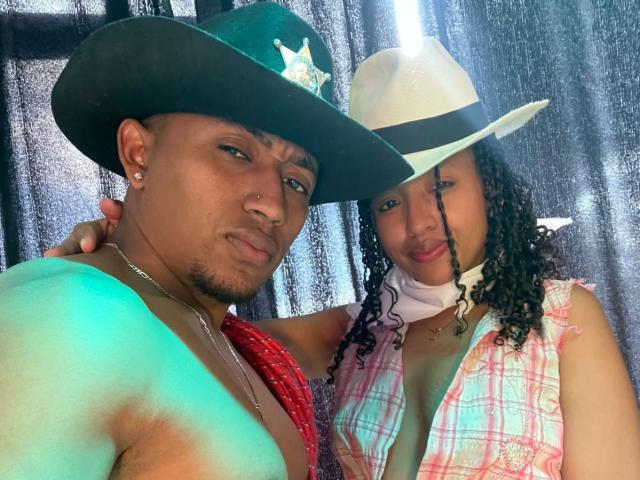 LilithAndAsmodeo with dark skin, wearing a green cowboy hat and red bandana, next to a woman with curly hair in a white hat and pink plaid vest.