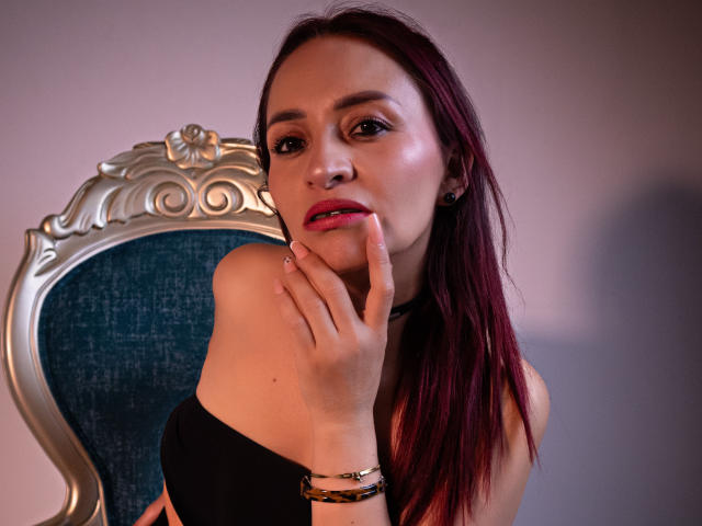 EmaCollins with red hair, black strapless top, touching her pink lips with her finger, wearing gold bracelets, sitting on a silver chair with blue upholstery.