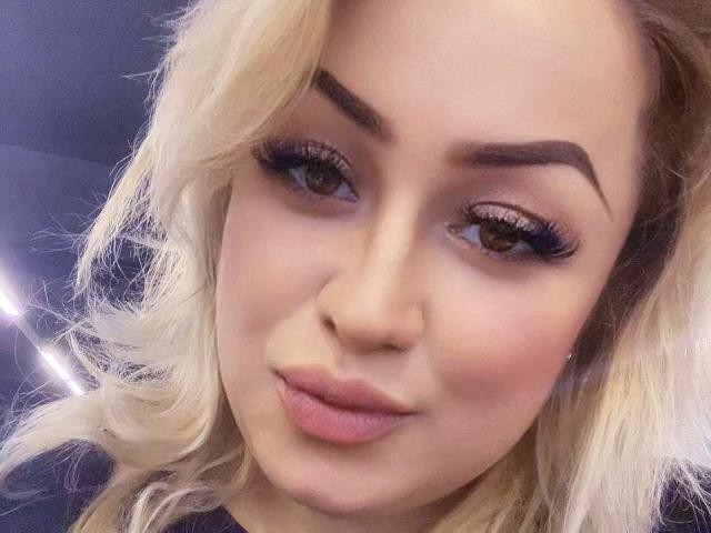Airisha a les cheveux blonds et ondulés, des sourcils épais et foncés, de longs cils volumineux et des lèvres roses légèrement entrouvertes. Elle regarde l'appareil photo avec un sourire subtil et confiant..