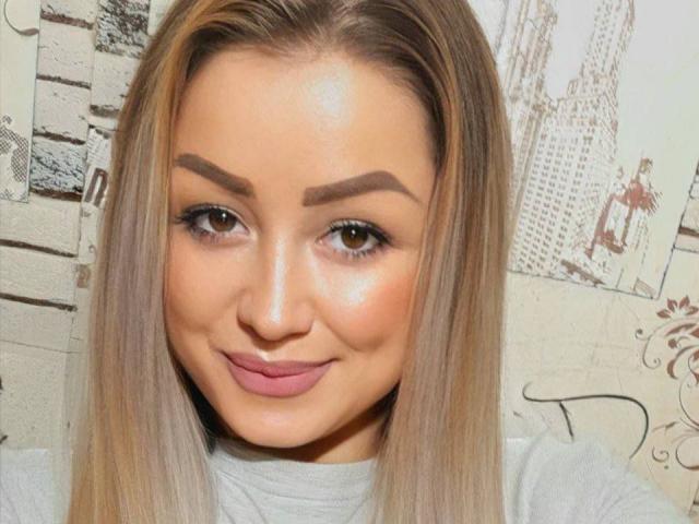 Airisha has smooth, tan skin, brown eyes, and thick eyebrows. She's smiling with pink lips, wearing a gray top, and has long, straight blonde hair.