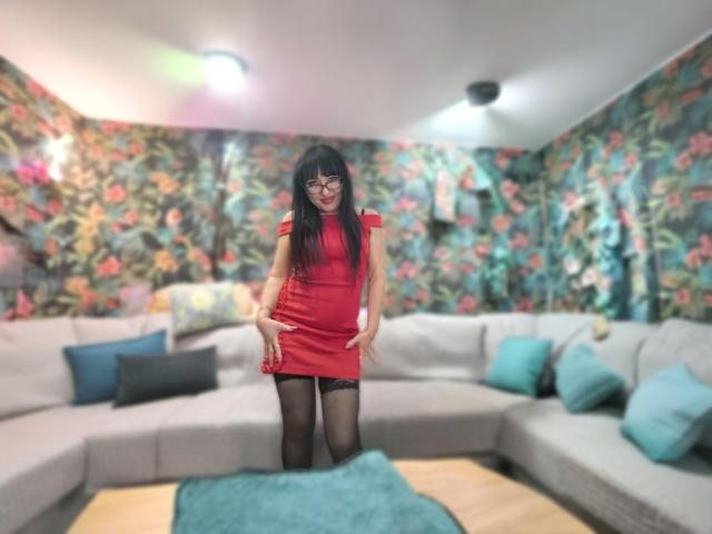 SusanLaNicex in a red dress, black stockings, and glasses, standing with her hands on her ass, in a colorful, floral-patterned room, on a gray sectional sofa.