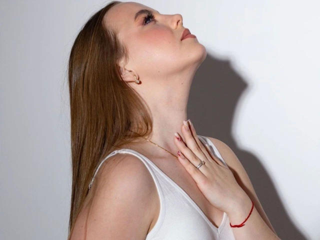 CherryMiss-atractiva, con cabello castaño largo, camiseta blanca, manos en el cuello, mirando hacia arriba, anillo rojo, aretes dorados, sombra en la pared blanca, piel clara, maquillaje sutil, anillo en el dedo..