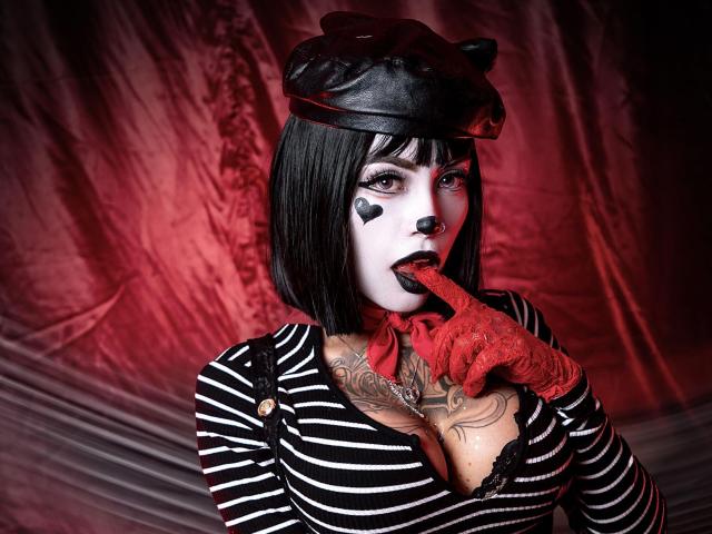 Sara Brooks, avec une coupe de cheveux courte noire, du maquillage blanc sur le visage, des motifs en forme de cœur, des gants rouges, un haut rayé, un soutien-gorge noir, un doigt dans la bouche, un chapeau noir, des tatouages, un fond rouge, séduisante, montrant ses seins..