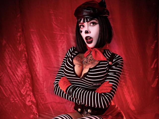 Sara Brooks met kort zwart kapsel, een kattenhoed, een gestreept shirt, een rode sjaal, een getatoeëerde borst, armen gekruist, een rode achtergrond, open mond, zwarte eyeliner, rode lipstick, gestreepte hoge sokken, met een geschokte blik..