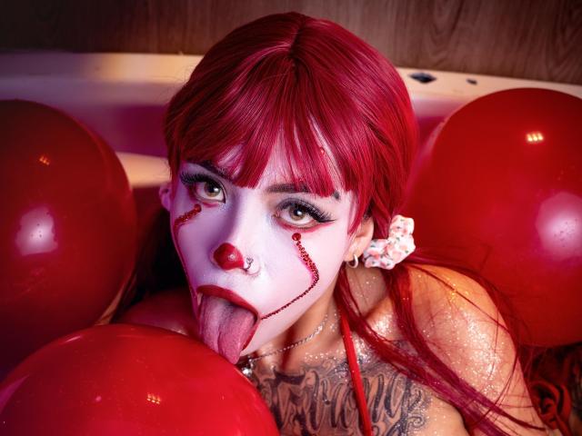 Sara Brooks, avec ses cheveux roux, son visage peint en blanc, son nez rouge et ses tatouages, sa langue tirée, entourée de ballons rouges, avec ses yeux verts et son rouge à lèvres rouge, dans un décor coloré et joyeux..