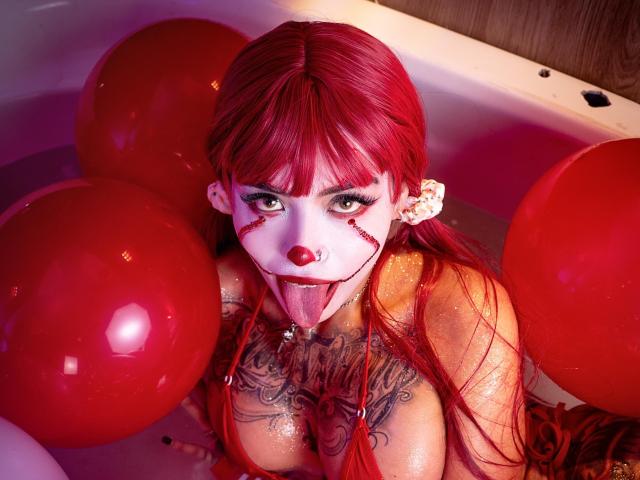 Sara Brooks, avec ses cheveux roux, son visage peint en blanc et son nez rouge, la langue tirée, dans une baignoire remplie de ballons rouges, montrant ses seins volumineux et sa poitrine tatouée, brillante d'eau..