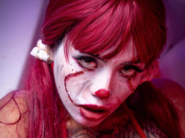 Sara Brooks, com cabelo ruivo, maquiagem branca no rosto, sangue nas bochechas e nariz vermelho, olhando intensamente com olhos verdes, usando um brinco branco, aparentando estar muito atraente..