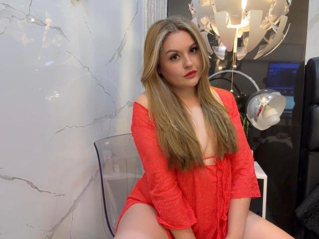 LaurenSpitz with long blonde hair, red off-shoulder dress, exposing her small tits and pussy, sitting on clear chair, looking at camera with red lipstick.