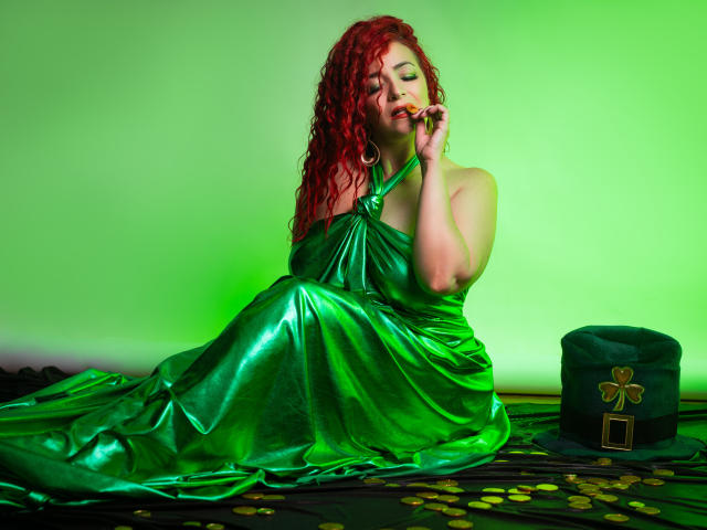 ScarlettthVega with red curly hair, green shiny dress, holding a cigarette, sitting on black with coins, green hat with gold button, green light, sexy, slim, small tits.