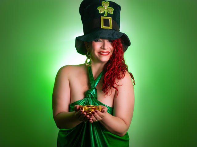 ScarlettVega with red curly hair, green dress, and top hat, holding gold coins, smiling, showing bare tits, wearing gold earrings, green glow, black hat with gold emblem.