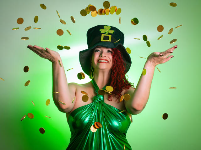 ScarletthVega stands center, arms raised, surrounded by falling gold coins. She wears a green metallic dress with a knot at the chest, and a black top hat with a gold shamrock. Her red curly hair contrasts with her fair skin. The green light behind her highlights her figure. The coins are mid-air, creating a dynamic sense of movement. ScarletthVega's joyful expression and bright red lipstick add a festive vibe. The composition emphasizes her celebratory pose and the coin cascade. The image is vibrant and playful.