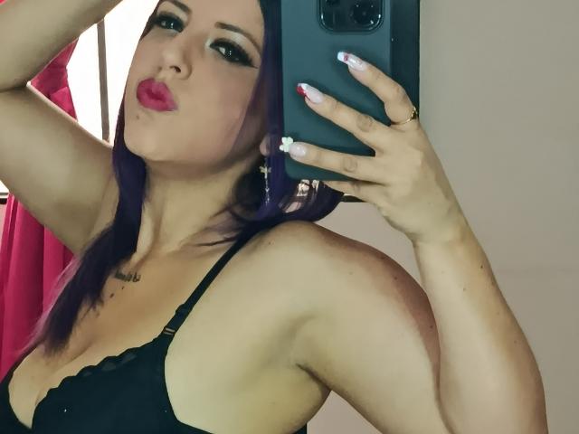 NicoleEmiller met paars haar, volle rode lippen, een zwarte bh, die een telefoon vasthoudt en haar borsten laat zien, nagels met witte tips, een sproetje in de oksel, een tatoeage onder de kin..