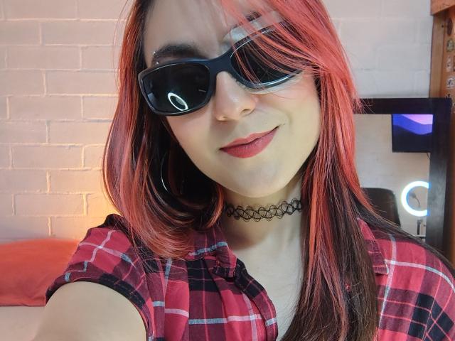 AmyStreetwise69 with red and black hair, black sunglasses, red plaid shirt, black choker, smiling, showing her lips, fair skin, and holding a selfie.
