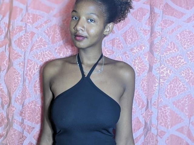 Ricka with dark skin, curly hair, small nose, wearing a black halter top, showing her tits, standing against a pink, floral-patterned curtain, looking slightly to the right.