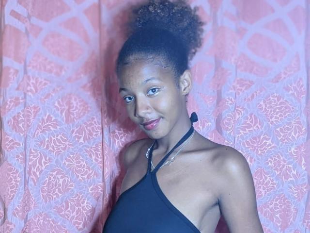 Ricka with dark skin, curly hair, and small tits, wearing a black halter top, smiling, standing against a pink patterned curtain. Her red lipstick is noticeable.