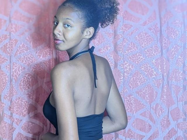 Ricka with curly hair, black halter top, showing back, small tits, looking over shoulder, smiling, pink floral-patterned curtain, blue light.