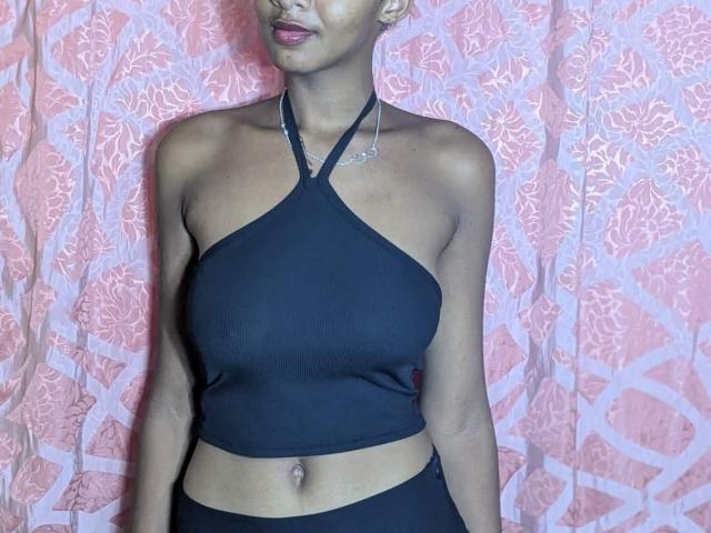 Ricka has dark skin, small tits, wearing a black halter crop top, black bottoms, and a silver necklace, standing against a pink patterned curtain.