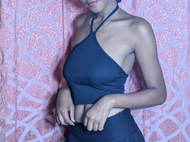 Ricka's small tits in a black halter top, exposing her midriff, with hands near her belly, wearing matching black bottoms, against a pink patterned curtain.