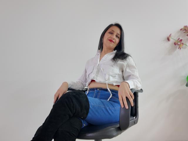 Cyntiia with black hair, red lipstick, white button-up shirt, blue jeans, and black thigh-high boots, sitting on a black chair, hands on her thighs, smiling.