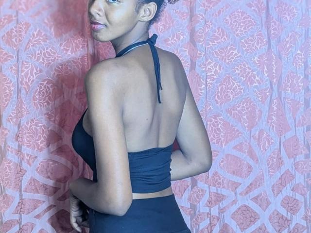 Ricka with dark skin, small to medium tits, black halter top, and black pants, standing with back to camera, looking over shoulder, smiling, short curly hair, pink floral curtain.