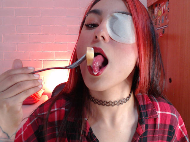 AmyStreetwise69 trägt ein rotes und schwarzes Karoshirt, eine schwarze Halskette, einen Augenklappe und hat eine rosa Zunge. Sie isst mit einer Gabel ein kleines Stück Käse und macht ein Selfie..