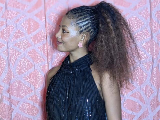 ClaraWish with braided ponytail, black sequined halter dress, pink flower earrings, smiling, looking to the side, curly brown hair, light brown skin, pink patterned background.