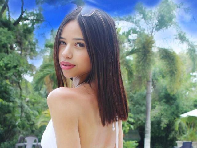 LiaBlom with medium brown bob, white halter top, pink lipstick, and small tits, looking over shoulder, blue sky, green trees, and palm trees in background, wearing sunglasses on head.