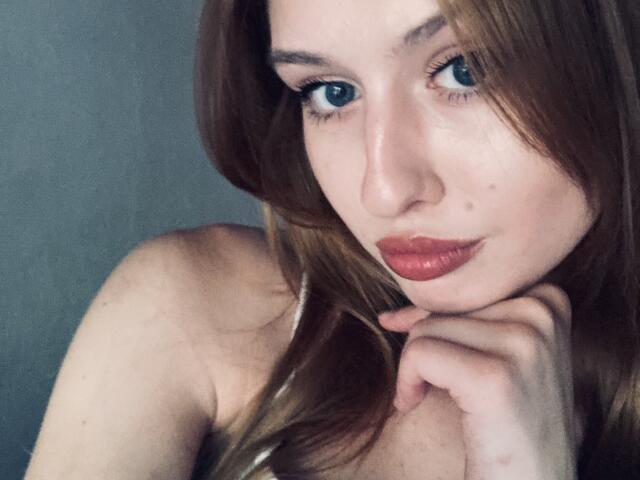 Nikollia, con la pelle chiara, gli occhi azzurri e il rossetto rosso, che tocca il mento con la mano destra, ha i capelli castani ed è senza top, mostrando le spalle scoperte..