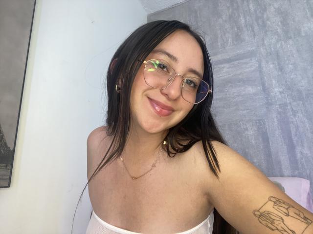 RosalieDupont with glasses, smiling, has small tits, black hair, tattoo on arm, white strapless top, gold necklace, and pink lips, in a selfie.
