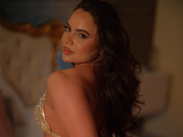 AshleeAngel, com cabelos castanhos ondulados, olhando por cima do ombro, vestindo um vestido dourado brilhante, mostrando suas costas e um pouco dos seios, em um quarto com pouca luz..