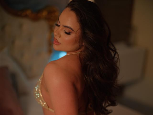 AshleeAngel, con cabello castaño ondulado, mirando hacia abajo, vestida con un sujetador dorado brillante que apenas cubre sus grandes y redondos senos. Una luz suave y cálida resalta su rostro sexy y sensual..