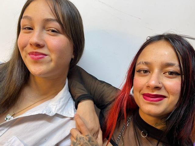 Roxxan e Emma, com cabelo castanho, batom rosa, camisa branca, de braços dadas, blusa de malha preta, cabelo ruivo-preto, batom rosa, piercing no nariz, tatuagem, sorrindo, com um fundo de parede branca..