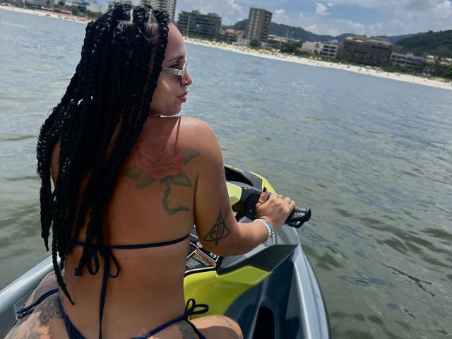 LunaX69, con trecce nere, indossa un bikini blu, ha un tatuaggio di una rosa sulla schiena e sta remando su un kayak giallo e nero, rivolta verso l'acqua, con una città sullo sfondo..