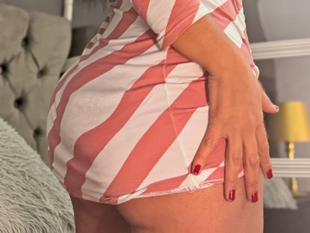 VeraCruz's butt in a pink and white striped dress, hand with red nails resting on it, showing her curvy ass and soft skin. Bedroom background.